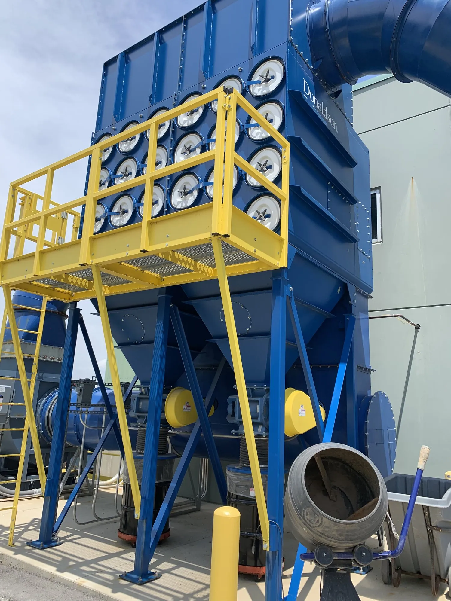 A blue Donaldson cartridge collector outdoors on structural steel, with a yellow access catwalk and ladder.