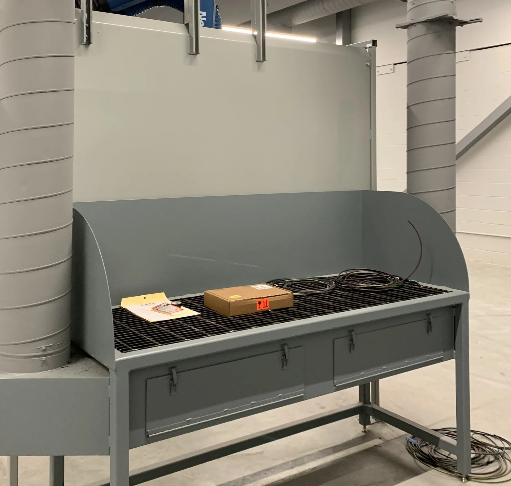A grey downdraft bench with a mesh work surface and back plenum, spiral ducting rising behind it to the roof.