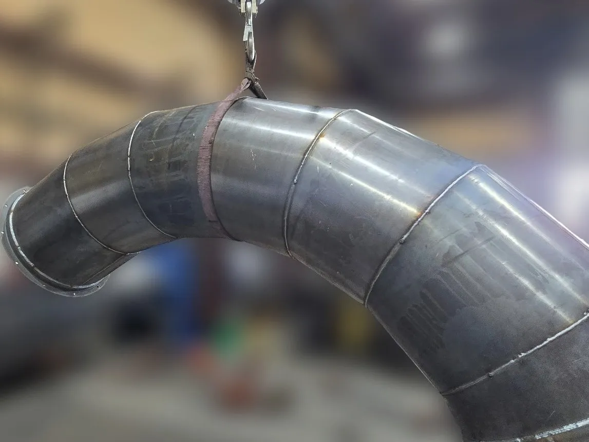 A heavy-gauge mitred steel duct elbow, built from welded segments, hanging from a shop crane.