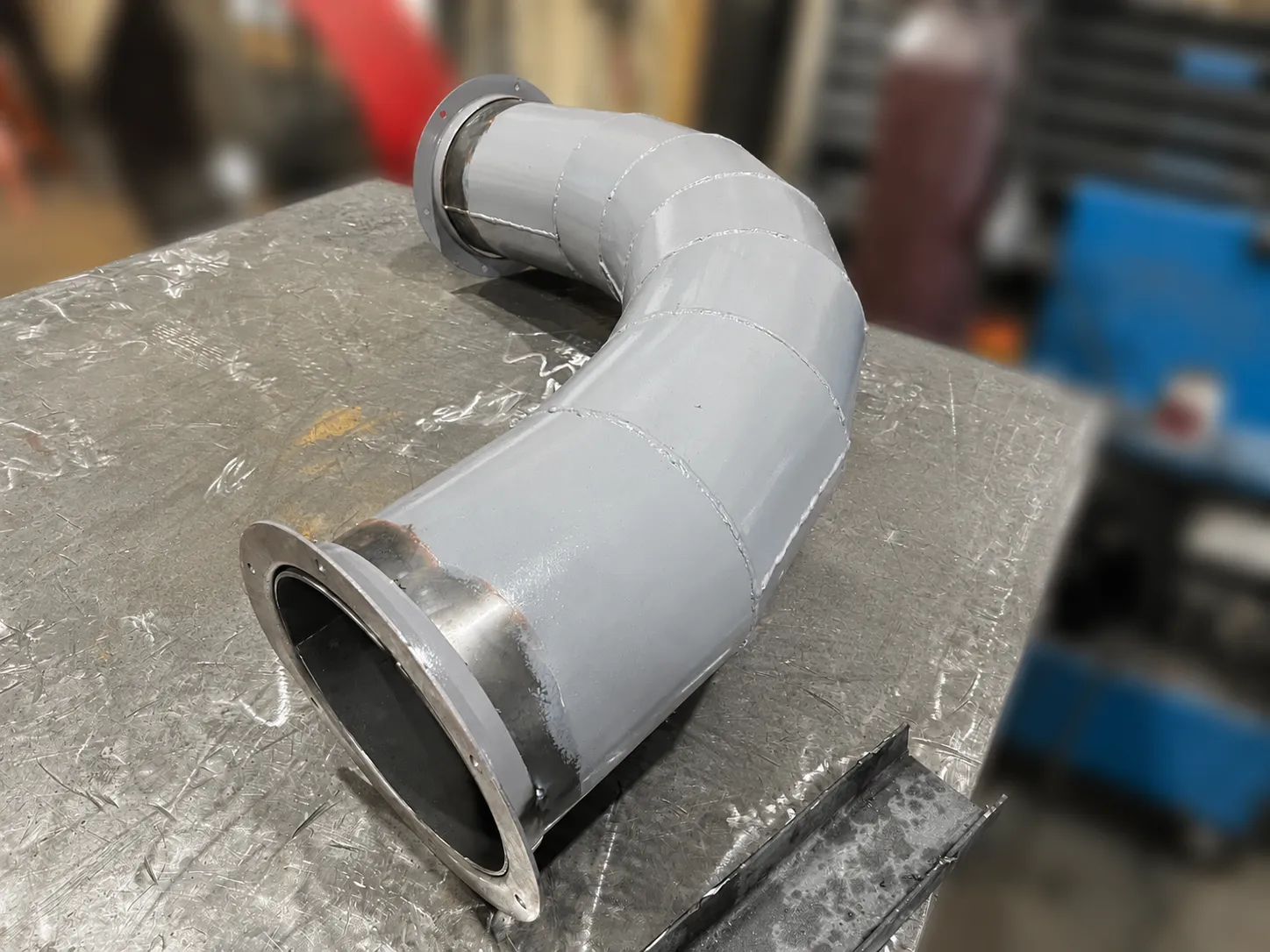 A fabricated duct elbow stepping down to a smaller flanged outlet, on a layout table.