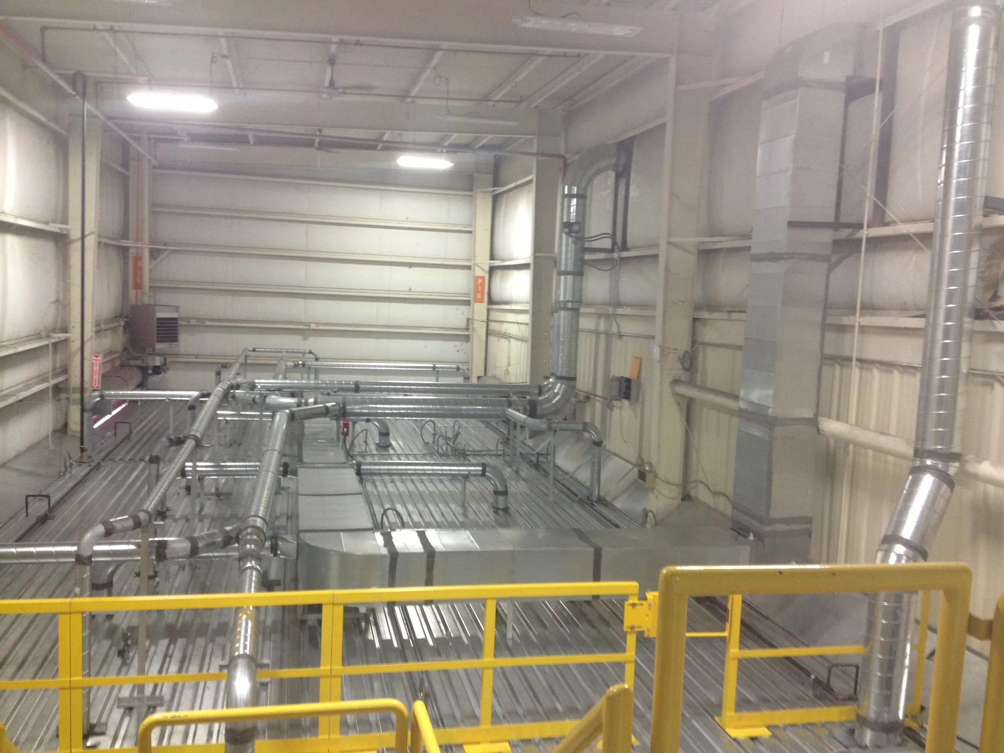 A run of spiral ductwork turning through a plant interior above a yellow guardrail.