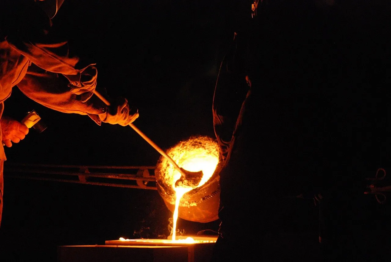 A foundry ladle tipping a single stream of molten iron into a mould, the pour lighting an otherwise dark shop floor.