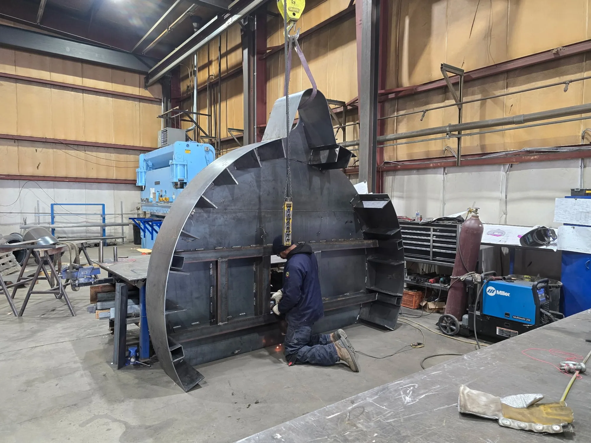A DCG fabricator kneeling to weld inside a large curved steel housing under a crane sling, in the company shop.