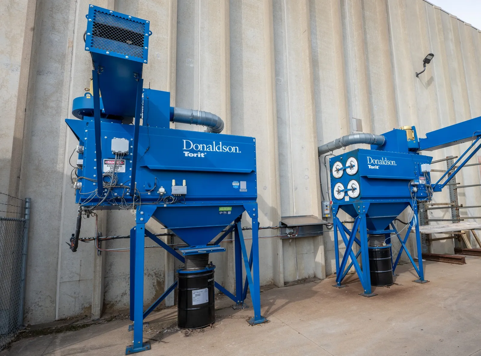 Two blue Donaldson Torit dust collectors on steel stands outside a plant wall, with collection drums beneath.