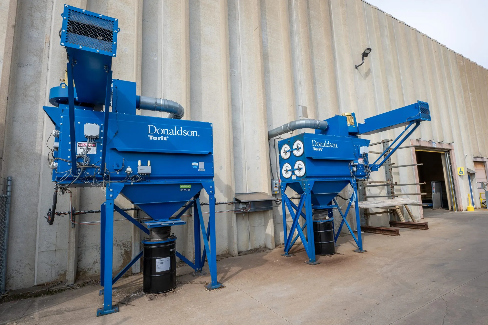 Two blue Donaldson Torit cartridge collectors on steel stands against a plant wall, collection drums beneath each.