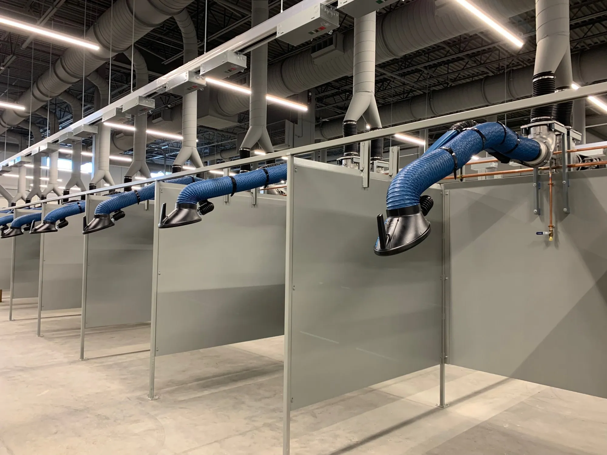 A row of welding booths in a training facility, each with an articulated source-capture fume arm dropping from an overhead duct run.