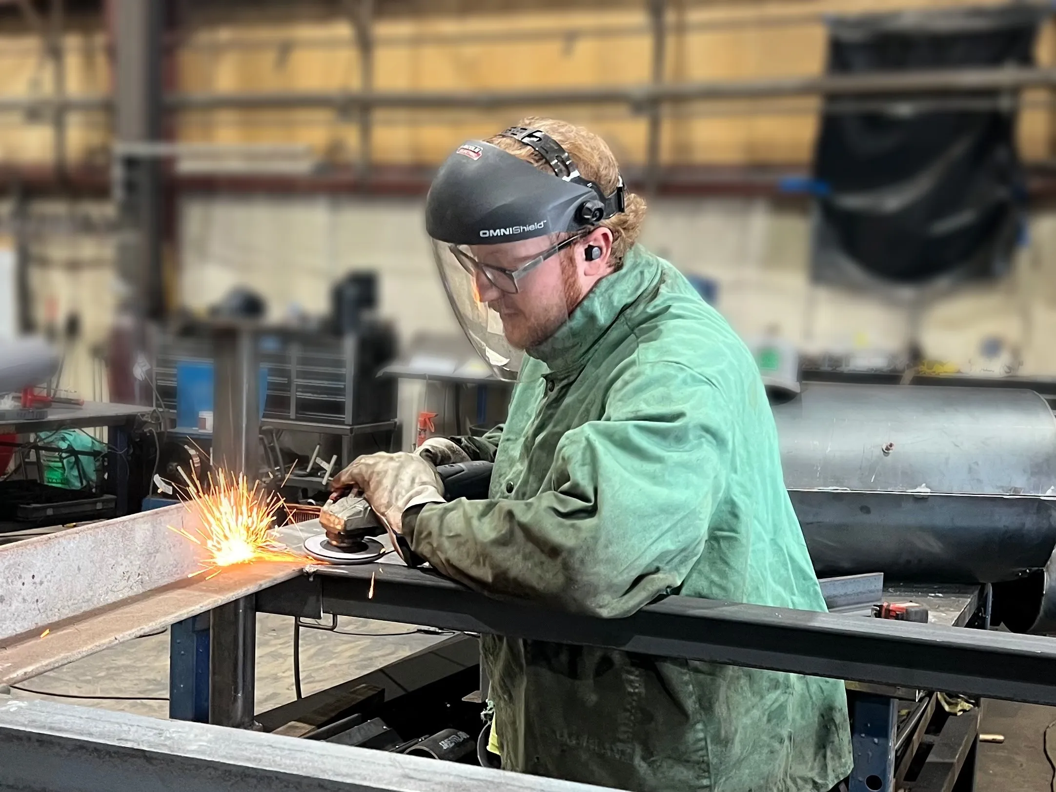 A DCG fabricator in a welding jacket and face shield grinding a steel beam, sparks flying.